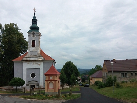 Snímek zachycuje kostel Nejsvětější Trojice v Hodňově, kde byl pokřtěn, a vpravo rodný dům čp. 14 - dochovala se jen přestavěná část někdejšího rozlehlého statku...