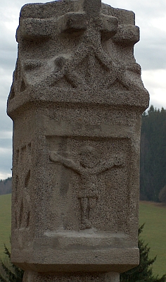 Boží muka s reliéfem Ukřižovaného, pocházející snad z roku 1521, zachycena v detailu a na pozadí jejího rodného domu, přestavěné zdejší školy