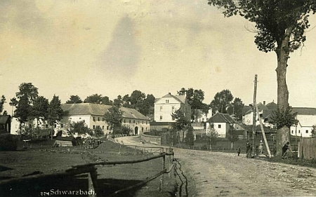 Stará pohlednice Černé v Pošumaví z fotoateliéru Josefa Wolfa zachycuje část obce u cesty na Český Krumlov - Proschkův rodný dům čp. 23 stál při ní o něco výše, na snímku vidět není